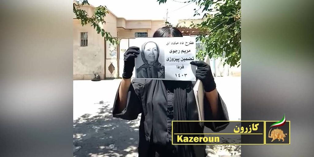 امرأة في كازرون تحمل في يدها لافتة كُتِب عليها: "برنامج المواد العشر لـ مريم رجوي ضمان لنصر الغد."